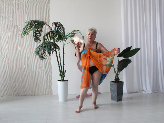 KayleSmoll, elderly with short white hair, holding an orange and blue sheer cloth over her black panties and bare ass, standing in front of two potted plants.