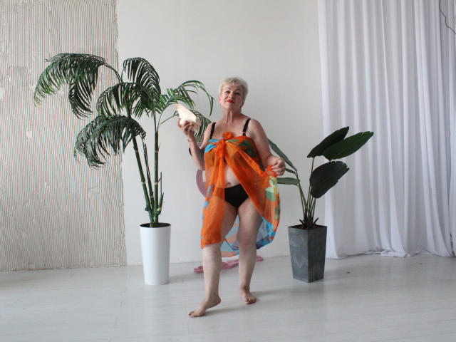 KayleSmoll with short white hair, black underwear, holding orange sheer fabric, standing barefoot, surrounded by green plants in white and gray pots, white floor, white curtain.