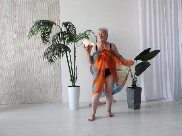 KayleSmoll, an older woman with short white hair, holds a large orange and blue scarf, revealing her bare black-tipped breasts and black panties, standing by two potted plants.
