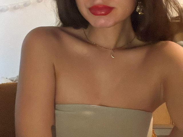AyanaLoveX with red lips, strapless grey top, bare shoulders, small tits, necklace, earrings, and a slight tan, in a soft light.