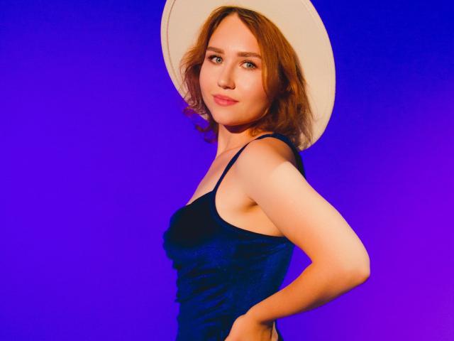 TeresaBirt with wavy brown hair, wearing a white hat and dark blue velvet dress, hands on her ass, looking at the camera with a subtle smile.