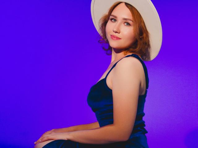 TeresaBirt has red hair, a white hat, and a dark blue dress, sitting with a gentle smile and soft eyes, against a vivid purple background.
