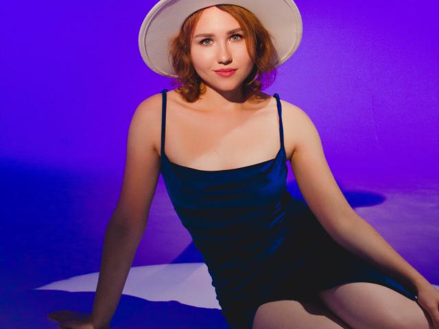 TeresaBirt with red hair, wearing a dark blue satin dress and white sun hat, sitting with her legs apart, showing her pale thighs, and a subtle smile.