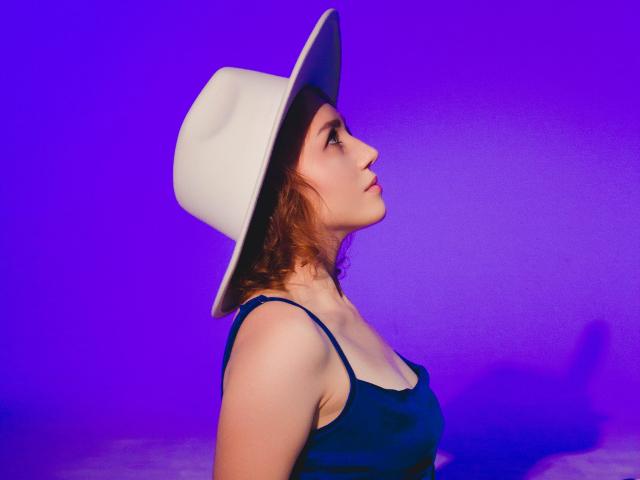TeresaBirt with short red hair, wearing a white hat and blue tank top, looking up with a slight smile, purple lighting, visible breasts, side profile, soft shadows.