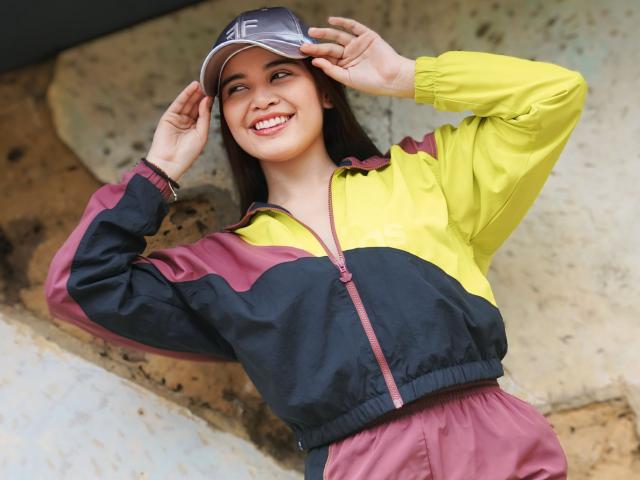 CamilaSofiia wearing a yellow and purple zip-up jacket, smiling with her hands on a gray baseball cap, brown hair, and a cute face, standing against a rough stone wall.