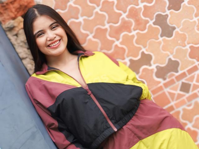 CamilaSofiia with long brown hair, smiling, wearing a yellow and maroon zip-up jacket, black chest panel, leaning against a blue surface, with a patterned brick wall behind her.