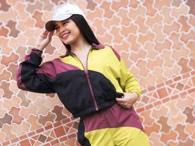 CamilaSofiia with dark hair, smiling, wearing a white cap, yellow and purple zip-up jacket, and yellow pants, touching her cap with her right hand.