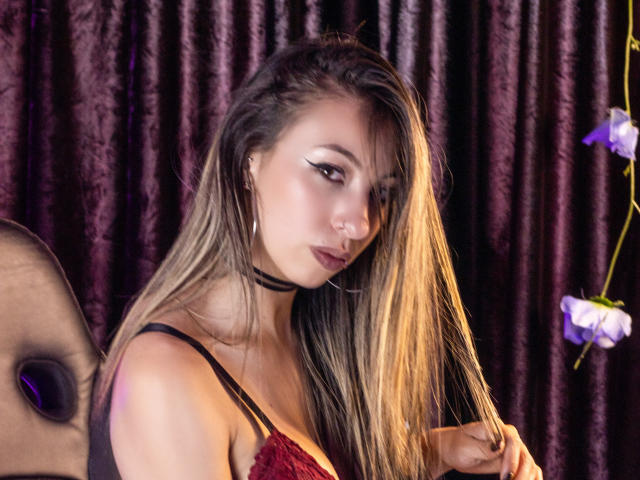 MilaBella with long blonde hair, black choker, red lace top, eyeliner, nose ring, and hoop earrings, holding her hair, showing her tits, and a slight pout.