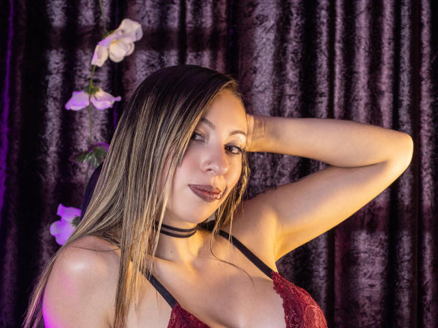 MilaBella with long blonde hair, wearing red lace bra, black choker, hand in hair, smiling, purple and pink flowers in background, soft lighting, sexy, attractive, medium tits.