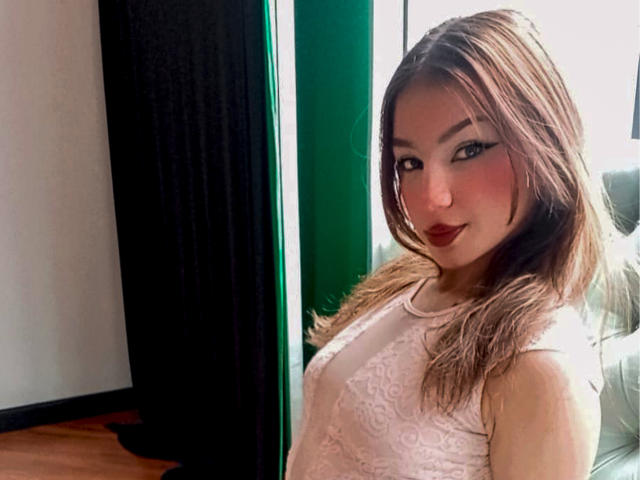 Ashley Miller, met bruin haar, draagt een witte top van kant en kijkt naar de camera met donkere lipstick en opvallende eyeliner. Ze staat bij een groene muur en een zwart gordijn..