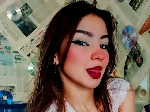 AshleyMiler has dark brown hair, white makeup, and red lipstick. She's looking sideways with black eyeliner, showing her small nose and wearing a white lace top.