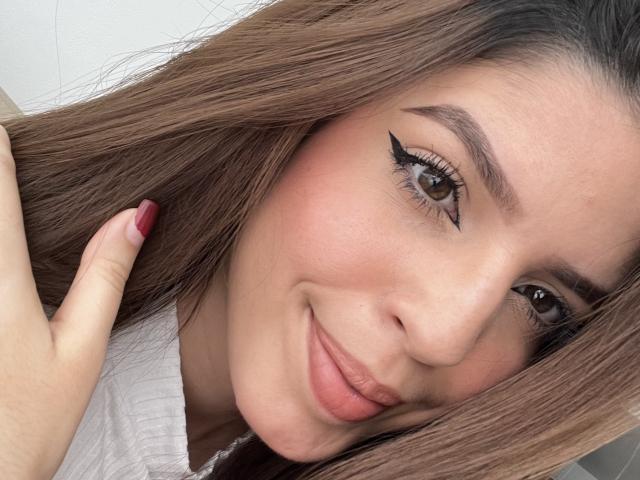 NatyBakers tiene ojos marrones, delineador de ojos alado y una ligera sonrisa. Su cabello castaño es largo, y lo está tocando con uñas pintadas de rojo. Lleva una blusa blanca..