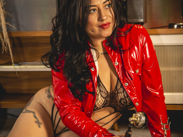 NoemiCami with long black hair, red shiny leather jacket, black fishnet lingerie, tattoos on thighs, sitting, looking at camera, red lipstick, sexy, confident, chest visible, nipples hidden.