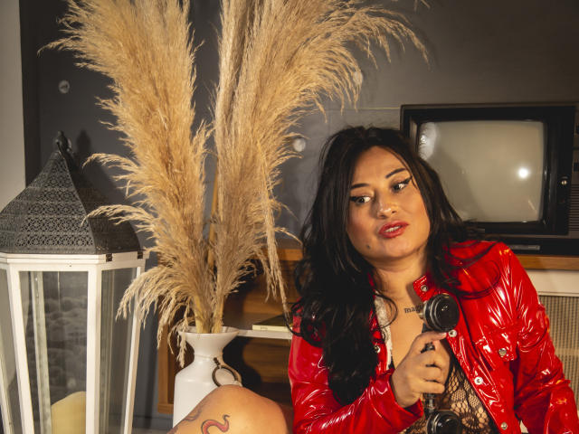 NoemiCami with dark hair, red shiny jacket, holding a camera, looking to the side, puffy ass tattoo, tall pampas grass, lantern, and old TV in the background.