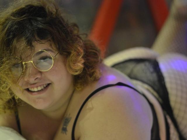 YviValentinny with curly brown hair, glasses, smiling, black lingerie, and fishnet stockings, showing her ass and a tattoo on her shoulder.