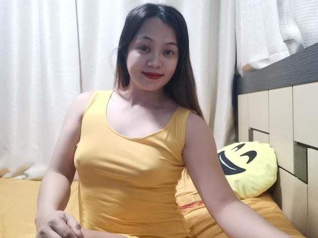 PinaylhianForU with long brown hair, yellow tank top, red lipstick, sitting on bed with yellow pillow, smiling, showing small tits, fair skin, in bedroom.