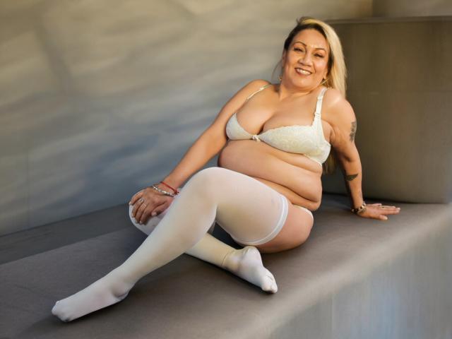 MatureYayita with white bra, white stockings, and blonde hair, sitting on gray bench, smiling, showing her tits, ass, and belly tattoo. Her pussy is covered by her bra.