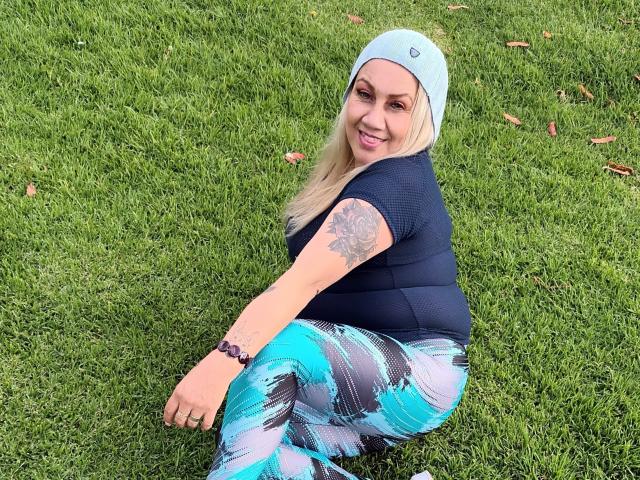 MatureYayita, blonde, wearing a light blue beanie, black top, and blue/white patterned leggings, sitting on grass, smiling, tattoo on right arm, bracelet on left wrist.