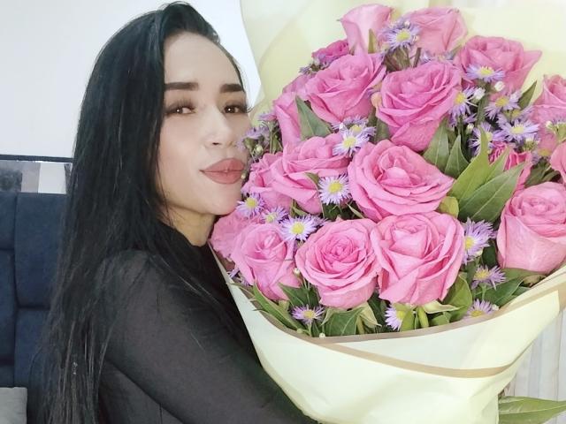 EvaMueller with long black hair, wearing a black top, holding a bouquet of pink roses and small purple flowers, smiling slightly, looking at camera.