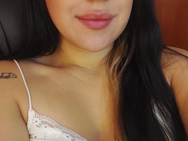 LauraRichis has a tattoo on her right shoulder, black straight hair with blonde highlights, and is wearing a white lace-trimmed bra, showing her small tits and pink lips.