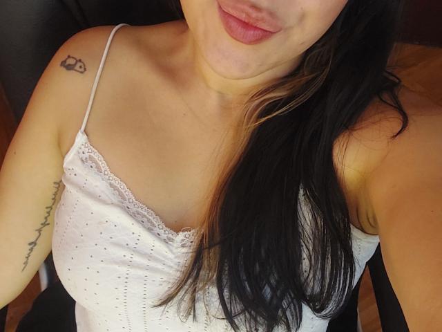LauraRichis with black hair, pouty pink lips, white lace-trimmed top, small tattoo on right shoulder, visible tattoo on left arm, showing her tits, seated.