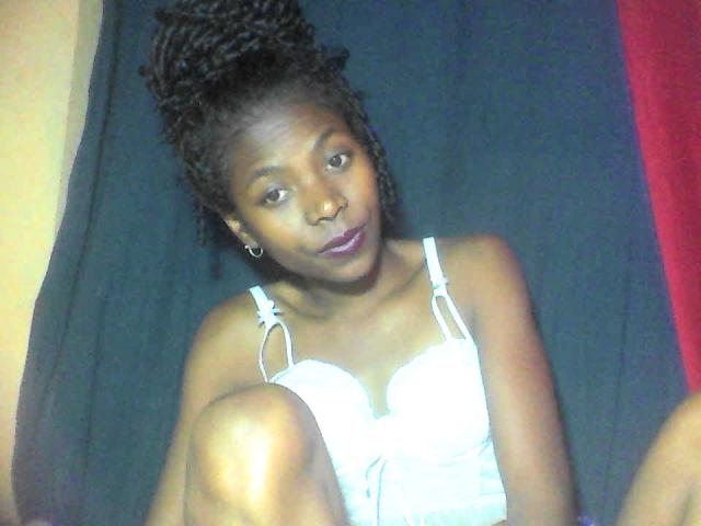 PrettyChocolat69, with dark skin and curly hair in a bun, wearing a white, strap-adorned, corset-like top, has her knee up and a slight, confident smile.
