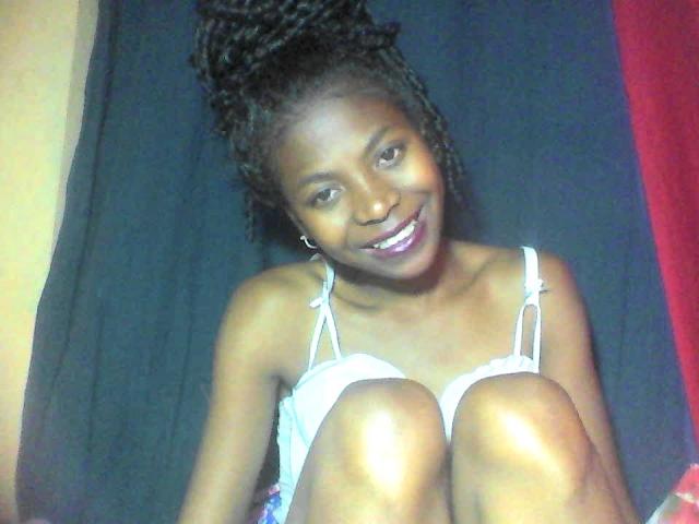 PrettyChocolat69 with dark skin, smiling, wearing a white spaghetti-strap top, knees up, black hair in a high ponytail, sitting against a black and red background.