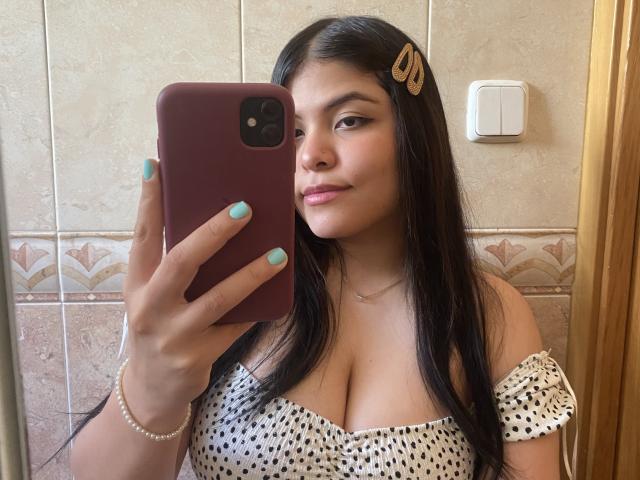 LuciaRoberts27 with medium tits, wearing a polka dot off-shoulder top, taking a mirror selfie with a pink phone, has long black hair, teal nails, and a gold hairclip.