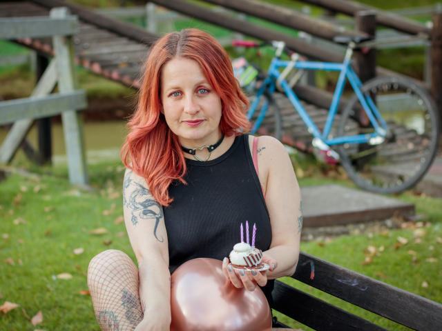 VioletaMora with red hair, black sleeveless top, and fishnet stockings, holding a cake with purple candles, tattoos on arm, blue bicycle in background, sitting on black bench.