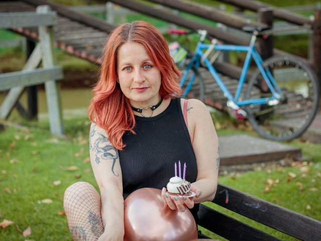 VioletaMora with red hair, black sleeveless top, tattoos, fishnet leg, holding cake with purple candles, sitting on bench, blue bike in background.