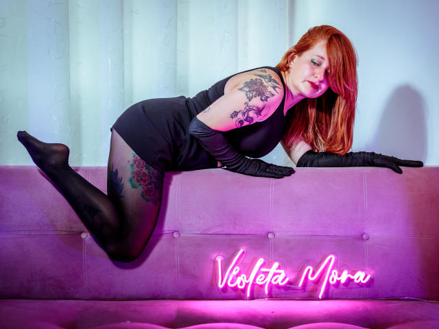 VioletaMora with red hair, black dress, and gloves, leans on a pink couch, showing tattoos on arm and thigh, wearing black stockings, her ass raised. Neon sign below.
