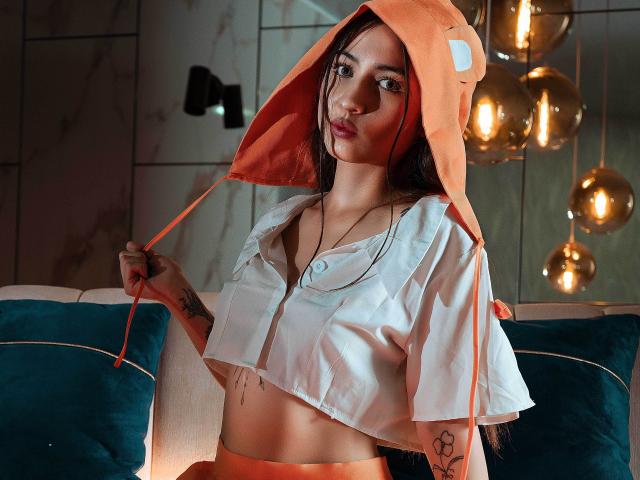 SopphiesWett with long dark hair, wearing a white crop top, orange hoodie with a cute animal face, and orange pants, holding hoodie strings, showing tattoos, looking sexy.