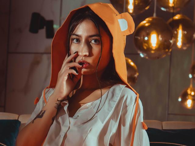SopphiesWett with brown hair, wearing an orange hood with bear ears, white shirt, biting finger, tattoo on wrist, and golden bubble lights in the background.