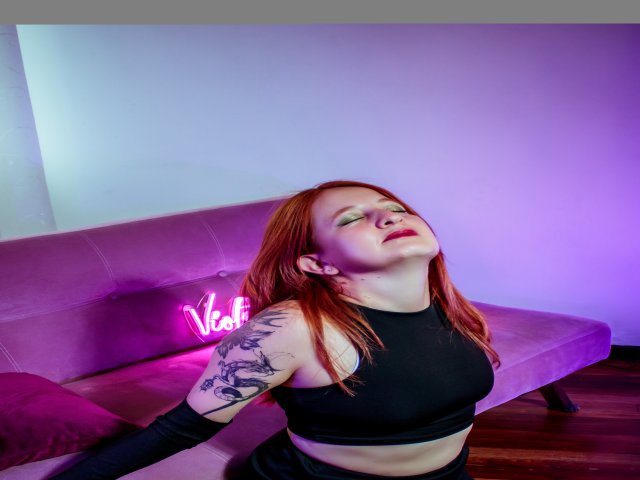 VioletaMora with red hair, eyes closed, head tilted back, wearing a black sports bra, showing a tattoo on her arm, in front of a purple sofa with neon sign.