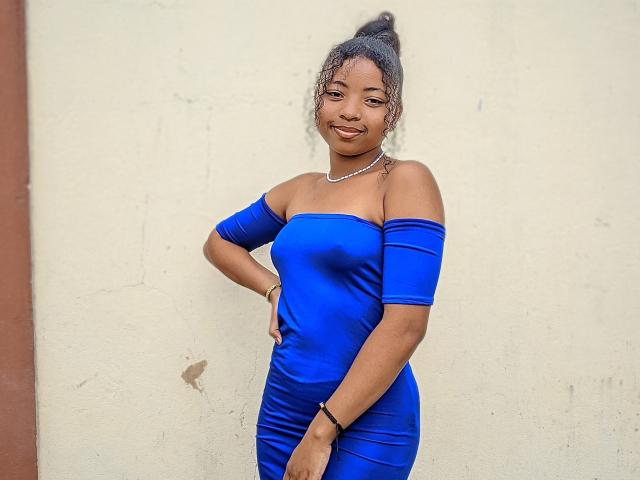 ShaniBelle, com pele escura, vestido azul com um ombro à mostra, cabelo grisalho encaracolado preso em um coque, sorrindo, com a mão na cintura, usando um colar e pulseira, em pé em frente a uma parede bege..