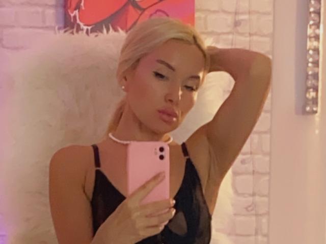 AvaGreenx with blonde hair, wearing black lingerie, holding pink phone, looking down, arm raised, white fluffy background, soft pink and white lighting.