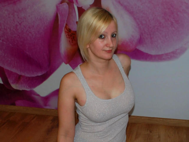 HotTitts-hot with blonde hair, wearing a sparkly gray tank top, showing small tits, standing in a room with a pink flower wall. Wooden floor, smiling, looking at camera.