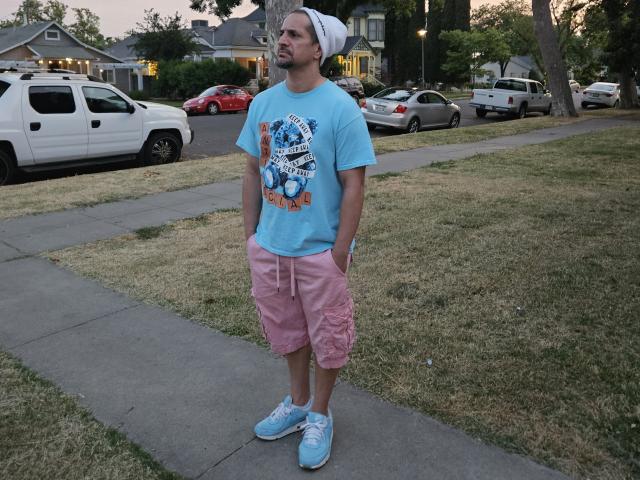SnackPants stands on a sidewalk, wearing a light blue shirt with a graphic, pink cargo shorts, blue sneakers, and a white cap, hands in pockets, looking off.