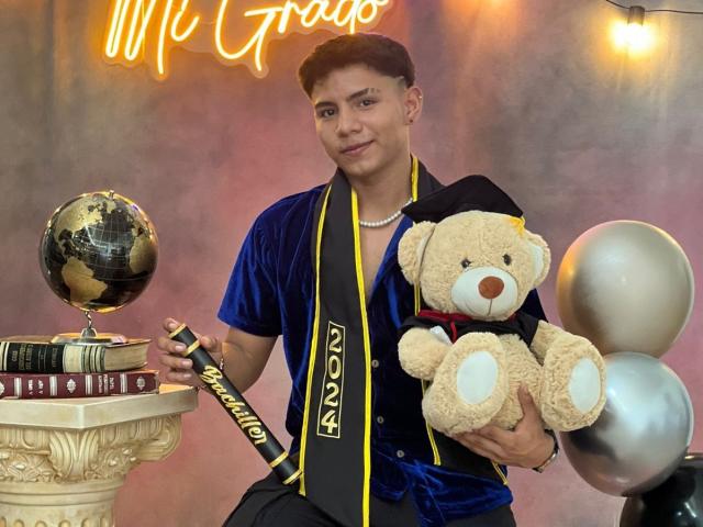 RichiMontes in a blue cap and gown, holding a teddy bear and diploma, with a globe and books on a pedestal, and silver balloons. He's wearing a yellow steeple.