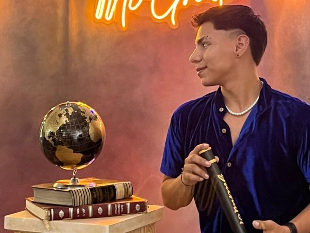 RichiMontes, con una camisa de terciopelo azul y un collar de perlas, sostiene una batuta negra mientras está de pie junto a un globo terráqueo y libros apilados. Un letrero de neón está encima..