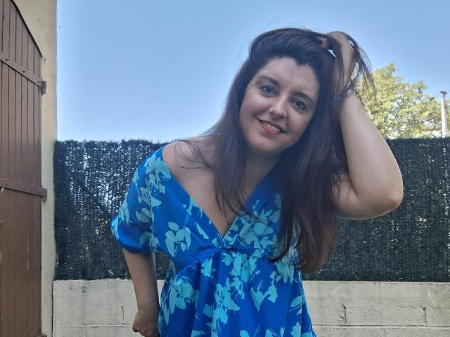 LaFrancaiseJessy, com cabelos castanhos longos, vestido floral azul, sorrindo, com a mão no cabelo, mostrando os seios, em pé em frente a uma cerca preta, céu azul claro e árvores verdes..