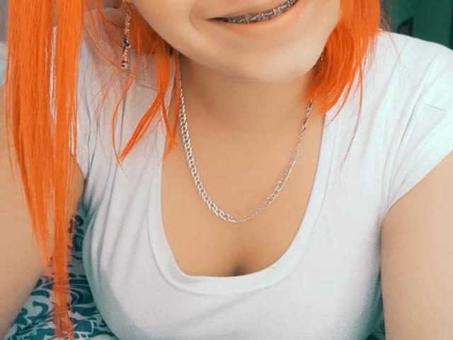 Kkatalina with bright orange hair, white shirt, silver chain necklace, and metal braces on her teeth, showing her cleavage. Her tits are visible, and she's smiling slightly.