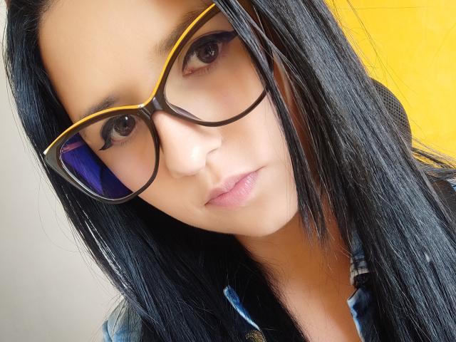 Kkatalina, con cabello negro y lacio, usa gafas con montura amarilla y una camisa de mezclilla azul. Tiene una expresión seria y un ligero tono rosado en los labios. Sus ojos son marrones y está mirando a la cámara..