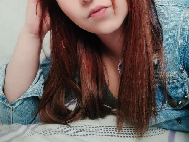 Kkatalina with long brown hair, wearing a blue denim jacket over a gray top, her hand in her hair, showing her pouty lips and a hint of her tits.
