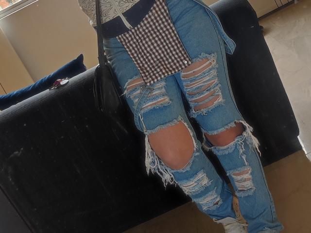 Kkatalina's ass is covered in ripped blue jeans with large holes showing her skin. She's wearing a checkered apron and white sneakers, standing next to a black couch.
