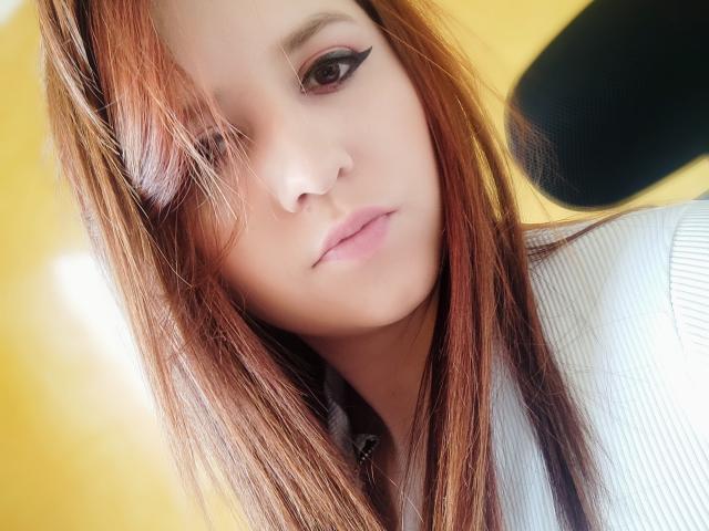 Kkatalina has light brown hair, black eyeliner, and pink lips. She's wearing a white ribbed shirt, with a black headrest in the upper right. Her expression is neutral.