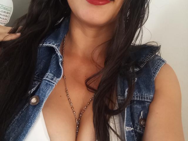 MadissonFontaine with dark hair, red lips, wearing a denim sleeveless jacket, white top, showing her big tits, and a chain necklace, holding a bottle in her right hand.