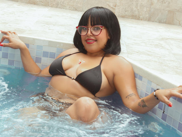 Morgan Moretz mit schwartem Bob, roten Brillen, einem schwarzen Bikini und rotem Lippenstift, sitzt im Pool, wobei Wasser ihre Brüste und ihren Hintern bedeckt. Sie hat ein Tattoo am rechten Arm und lächelt..