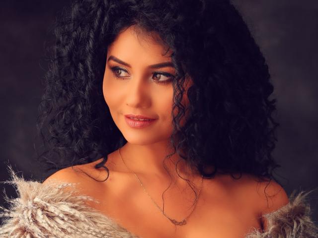 CanelaLebrad with curly black hair, tan skin, brown eyes, and a small smile, wearing a furry off-shoulder coat and a delicate necklace, looking slightly to the side.