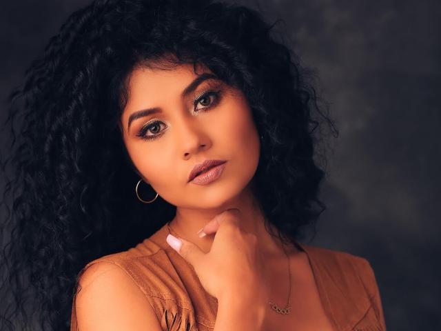 CanelaLebrad with curly black hair, tan skin, brown eyes, and hoop earrings, wearing a tan top, touching her chin, looking seductive with pink lipstick.